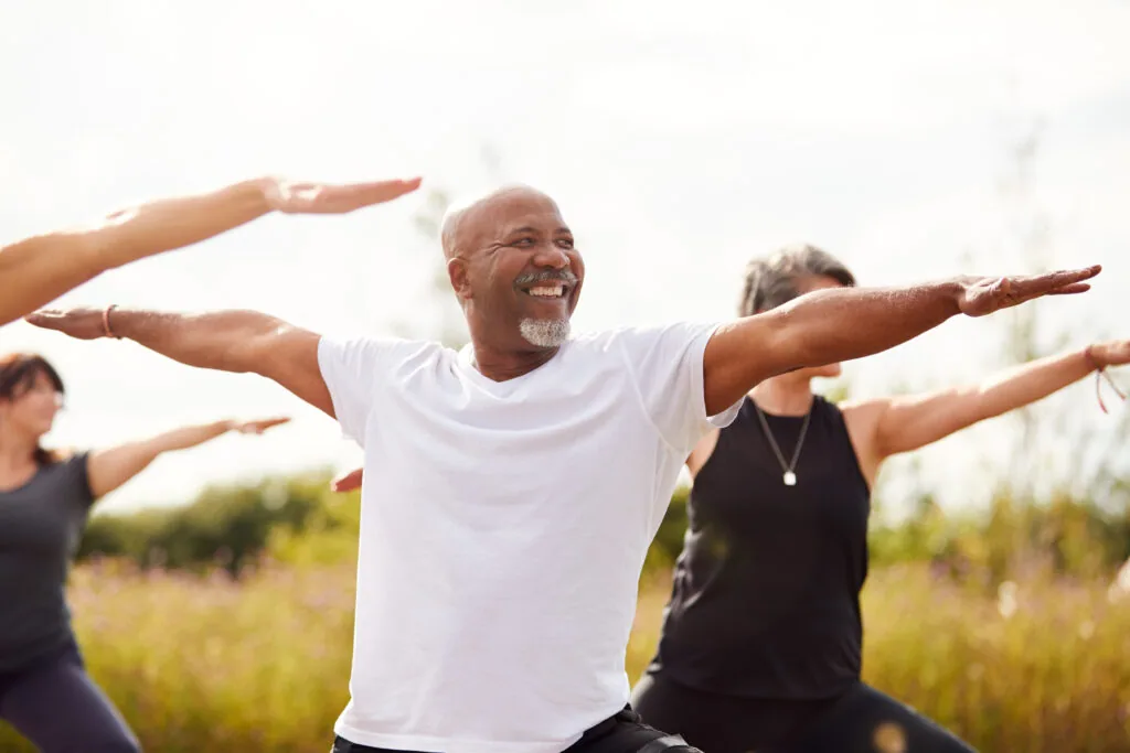 Man in white shirt smiling with arms extended during outdoor group exercise class with other participants