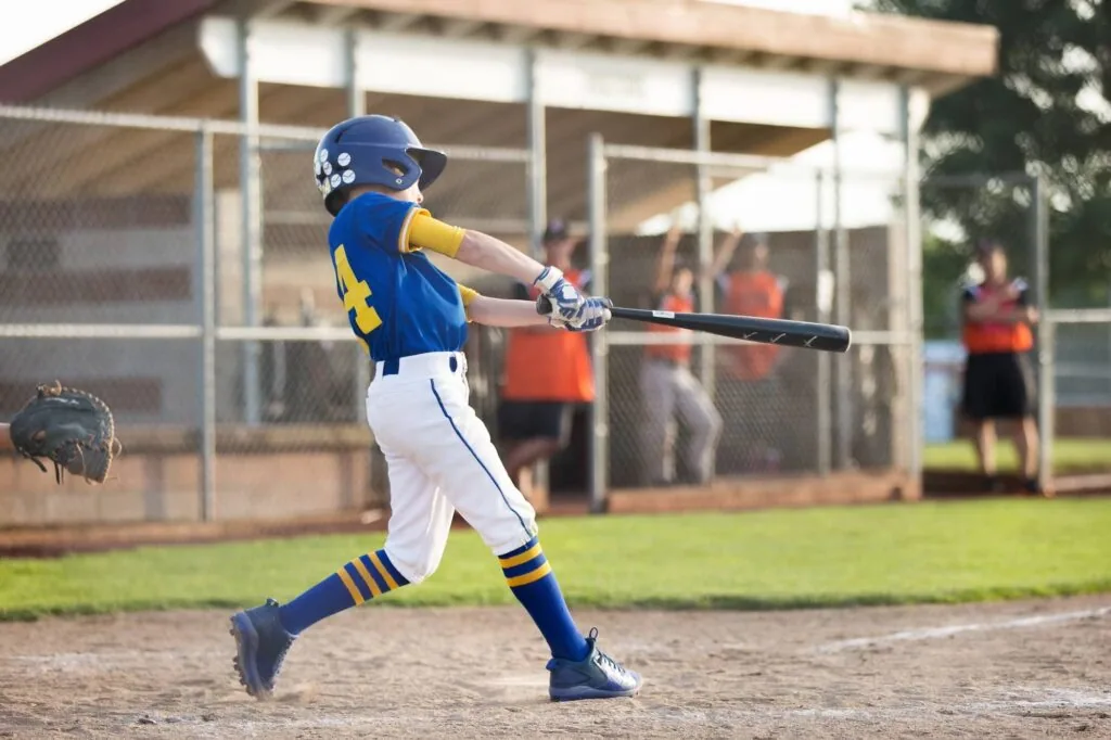 Young baseball player in blue and yellow uniform swinging bat during game at field with grandstand in background