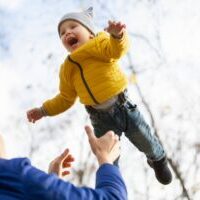 Toddler in yellow jacket and white hat being tossed in the air by adult in blue jacket, with snowy background